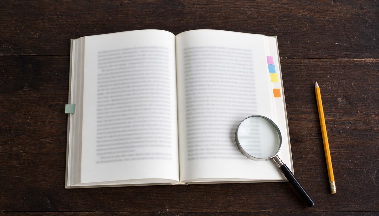 어두운 나무 책상에 책 한 권이 펼쳐져 있고 오른쪽 페이지 위 돋보기와 연필, 색깔 탭 다섯 개가 놓여 있다.