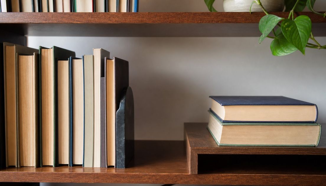 Upright plain books rest against a dark metal bookend beside an open gap, while two large clothbound books lie flat on a fully supporting shelf.