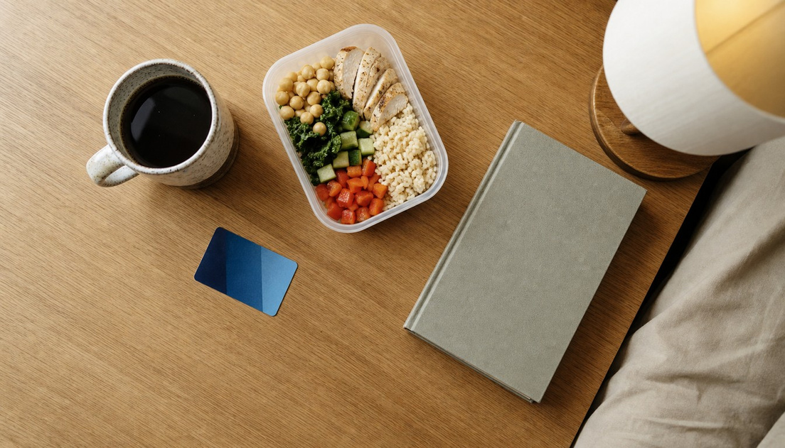 A closed gray book lies on a pale wooden table with a coffee mug, lunch container, plain blue card, and lit lamp.